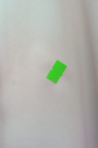 Mały zielony kawałek taśmy spoczywa na jednolitym, jasnym tle z tkaniny, przypominającym materiały często spotykane w kolekcjach marki Sukienka Bokserka.