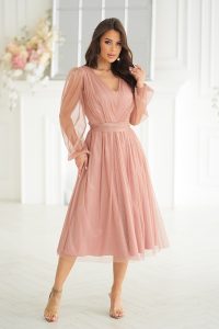 Kobieta w jasnoróżowej sukience midi z długim rękawem pozuje w jasnym, eleganckim pomieszczeniu z kwiatowym wystrojem. Subtelne detale z brokatowej siatki dodają odrobinę blasku do wyrafinowanego wyglądu.