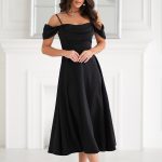 Kobieta w czarnej Sukience Midi 325-06 z opadającymi ramiączkami i szpilkach stojąca w jasnym, eleganckim pomieszczeniu.