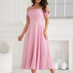 Kobieta w eleganckiej różowej sukience midi, z opadającymi ramiączkami, stoi w pomieszczeniu przy białej ścianie i szklanych butelkach.