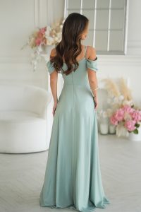 Kobieta w jasnoniebieskiej sukience z drapowaniem stoi odwrócona tyłem w jasnym, eleganckim pokoju wypełnionym różowymi i białymi kwiatami.