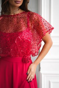 Kobieta w eleganckiej sukience na ramiączkach, ozdobionej czerwonymi cekinami, przezroczystą błyszczącą peleryną i kwiatowym detalem w talii.