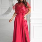 Kobieta w długiej, jasnoczerwonej Eleganckiej Sukience z koronkową górą i rozciętą spódnicą, stojąca w białym eleganckim pokoju.