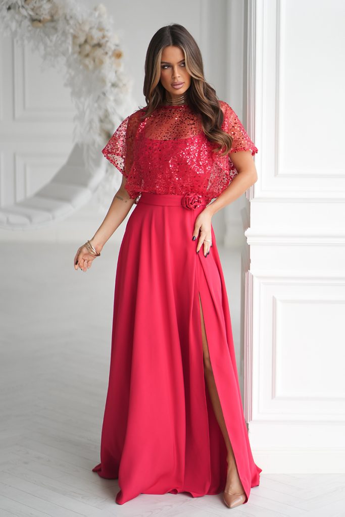 Kobieta w długiej, jasnoczerwonej Eleganckiej Sukience z koronkową górą i rozciętą spódnicą, stojąca w białym eleganckim pokoju.