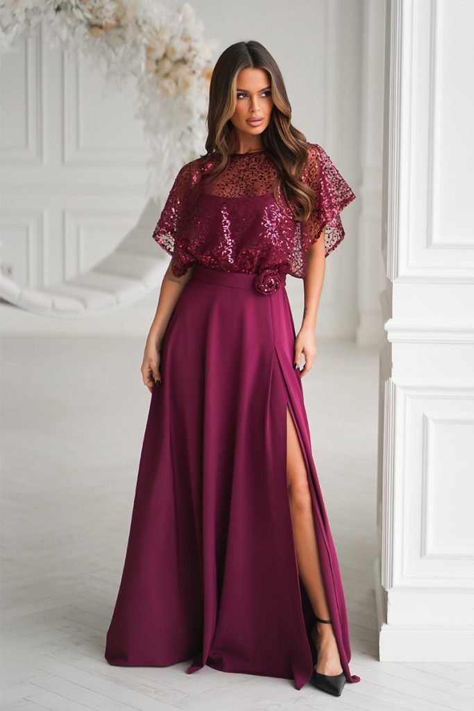 Elegancka sukienka na ramiączkach z cekinowym topem i wysokim rozcięciem, którą nosi kobieta stojąca w eleganckim, białym wnętrzu.