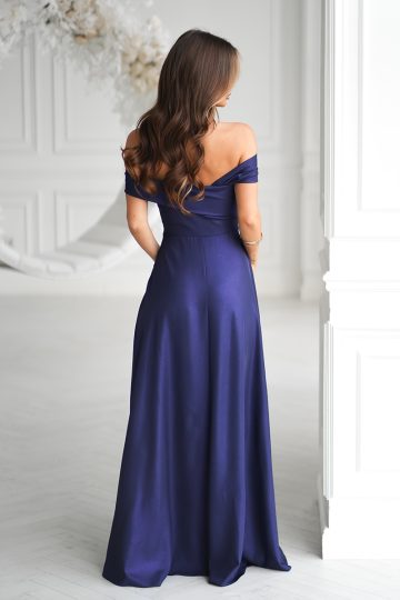 Kobieta w Długiej Sukni Wieczorowej o metalicznym połysku, stojąca tyłem do kamery w białym pokoju, prezentuje ponadczasową elegancję w granatowej sukni z odkrytymi ramionami, sięgającej podłogi.