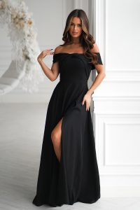 Kobieta w długiej sukni wieczorowej z dekoltem odkrywającym ramiona i rozcięciem do uda, stojąca w białym eleganckim pokoju.