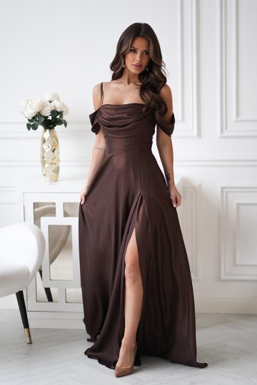 Kobieta w eleganckiej sukience z drapowaniem, z brązową suknią bez ramion z rozcięciem, stoi w pomieszczeniu przy białym stole ozdobionym kwiatami.