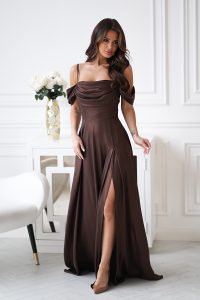 Kobieta w brązowej eleganckiej sukience z drapowaniem i rozcięciem na wysokości uda, stojąca w eleganckim białym pokoju.