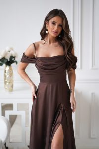 Kobieta w eleganckiej sukience z drapowaniem, stojąca w pomieszczeniu, jej falujące włosy i złote kolczyki łapią światło w pobliżu białego stołu.