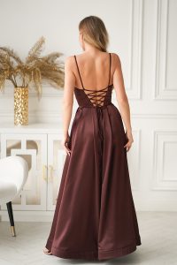 Kobieta w bordowej, połyskującej sukni wieczorowej z gorsetowym sznurowaniem stoi odwrócona tyłem w stylowym, eleganckim pomieszczeniu.