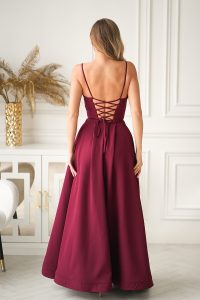 Kobieta w bordowej Długiej Sukni Wieczorowej z gorsetowym sznurowaniem stoi w eleganckim, białym pokoju ozdobionym złotym dekorem.