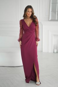 Kobieta w ciemnoróżowej sukni z przezroczystymi rękawami z cekinów, stojąca w jasnym, eleganckim pomieszczeniu.