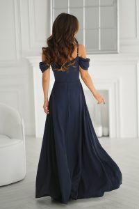 Kobieta w eleganckiej, zwiewnej granatowej sukience stoi w jasnym, białym pokoju, odwrócona plecami do aparatu.