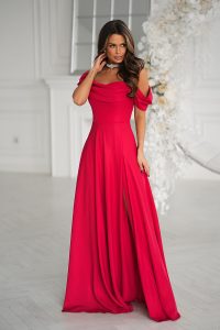Kobieta w powiewnej czerwonej sukni z odkrytymi ramionami i rozcięciem sięgającym do połowy uda, stojąca w eleganckim białym pokoju.