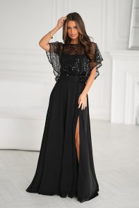 Kobieta w czarnej, sięgającej podłogi sukni wieczorowej z prześwitującym gorsetem pokrytym cekinami, stojąca w jasnym, eleganckim pomieszczeniu.