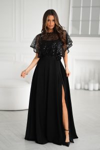 Kobieta w długiej, czarnej sukni wieczorowej z cekinowym dekoltem i rozcięciem z boku, stojąca w jasnym, eleganckim pomieszczeniu.