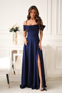 Kobieta w granatowej sukni z odkrytymi ramionami i rozcięciem sięgającym do połowy uda, stojąca w eleganckim białym pokoju.