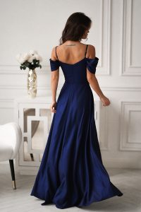 Kobieta w eleganckiej granatowej sukni z odkrytymi ramionami, stojąca w białym, stylowo urządzonym pokoju.