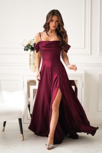 Kobieta w bordowej sukni z odkrytymi ramionami i rozcięciem sięgającym do połowy uda, stojąca w eleganckim białym pokoju.