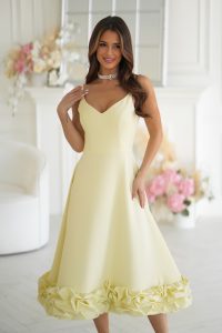 Kobieta w żółtej sukience midi z marszczonym dołem stoi w jasnym pokoju ozdobionym kwiatowymi dekoracjami, promieniując niewymuszoną elegancją.