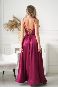 Kobieta w długiej sukni w kolorze głębokiego magenty, z wiązaniem z tyłu, stojąca w eleganckim, białym pokoju.