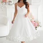 Kobieta w sukience midi ecru z kwiatowym wykończeniem, stojąca z gracją w eleganckim pokoju ozdobionym świeżymi kwiatami.