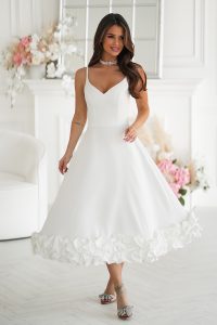 Kobieta w sukience midi ecru z kwiatowym wykończeniem, stojąca z gracją w eleganckim pokoju ozdobionym świeżymi kwiatami.