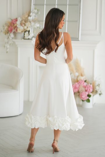 Kobieta w Sukience Ecru z marszczonym dołem stoi w pomieszczeniu, odwrócona tyłem, w pobliżu kwiatowych dekoracji i białego krzesła, prezentując idealną Sukienkę Midi na każdą okazję.