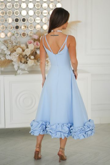 Kobieta w Błękitnej Sukience Midi z marszczonym dołem i kwiatowym wykończeniem stoi w pomieszczeniu, odwrócona tyłem do kamery.