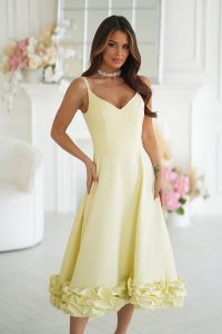Kobieta w efektownej żółtej sukience midi z marszczonym dołem, stojąca z gracją w jasnym, eleganckim pokoju ozdobionym kwiatami.