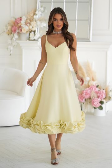 Kobieta w efektownej sukience - żółta sukienka midi z kwiatowym wykończeniem - stoi z gracją w białym pokoju ozdobionym kwiatami i lustrem.