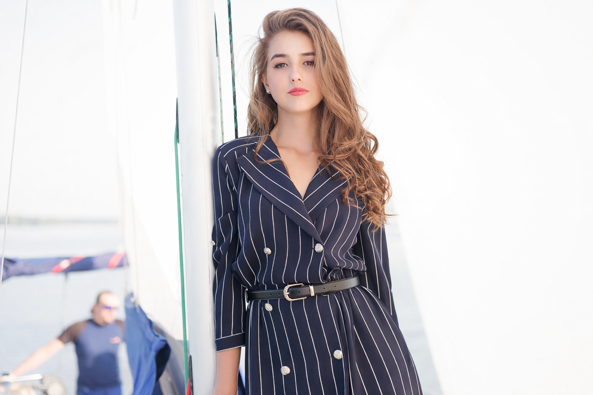 Kobieta w marynarskich sukienkach w prążki stoi na żaglówce; żagle i woda są widoczne w tle, oferując inspirację jak nosić eleganckie stylizacje na co dzień.