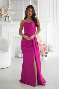 Kobieta w fuksjowej sukni bez ramiączek z rozcięciem na wysokości uda stoi w eleganckim białym pokoju z kwiatowym wystrojem.