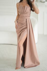Kobieta w eleganckiej, bez ramiączek, pudrowo-różowej sukni z wysokim rozcięciem, stojąca w pomieszczeniu na jasnej podłodze.