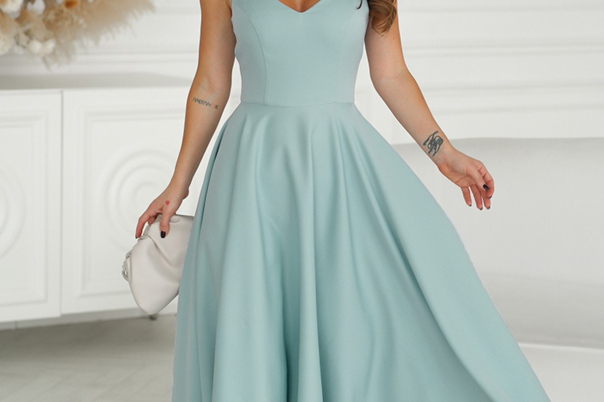 Kobieta w jasnoniebieskiej sukience bez rękawów z kolekcji butik 2026 marki kolory sukienek, trzymająca białą kopertówkę i stojąca w jasnym, eleganckim pomieszczeniu.