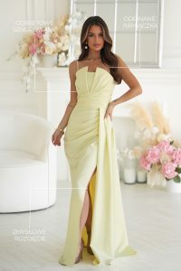 Kobieta w jasnożółtej sukni z geometrycznym dekoltem i eleganckim rozcięciem, stojąca w pomieszczeniu obok pastelowych kwiatów; oznaczone są detale konstrukcyjne sukni, takie jak odpinane ramiączka i wyraziste linie.