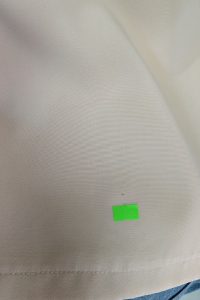 Biała tkanina z małą, jasnozieloną prostokątną naklejką przy dolnej krawędzi, przypominająca wzory spotykane w kolekcjach outletowych, takie jak beżowy kombinezon z zakrytymi ramionami.
