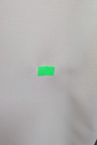 Mały kawałek jasnozielonej taśmy przyklejony do gładkiej, jasnej powierzchni, przypominający delikatny odcień charakterystyczny dla beżowego kombinezonu.