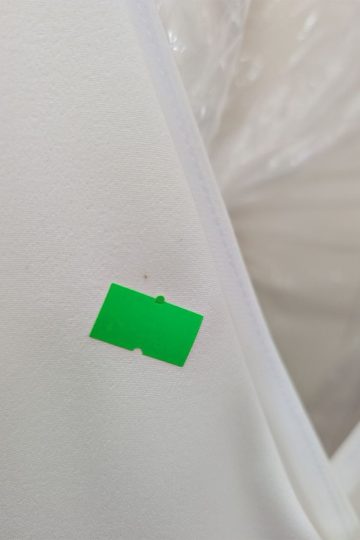 Mała zielona naklejka na białej, teksturowanej powierzchni z szwem i przezroczystym plastikiem w tle, przywodząca na myśl wyrafinowaną estetykę charakterystyczną dla „Eleganckiej Dopasowanej Sukienki”.