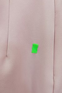 Mały kawałek zielonej taśmy przyklejony jest do jasnoróżowej tkaniny, a po lewej stronie widać szew, co sugeruje, że może to być fragment różowej sukienki.