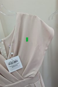Jasnobeżowa sukienka na wieszaku z zieloną naklejką i białą metką z ceną przymocowaną w pobliżu zamka błyskawicznego, przypominająca beżowy kombinezon, idealna na zakupy w outletach.