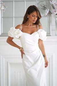 Kobieta w eleganckiej białej sukience z bufiastymi rękawami, stojąca w pomieszczeniu przy białej ścianie i lustrze.