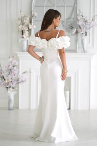 Kobieta w eleganckiej białej sukni z odkrytymi ramionami stoi w jasnym pomieszczeniu ozdobionym kompozycjami kwiatowymi.