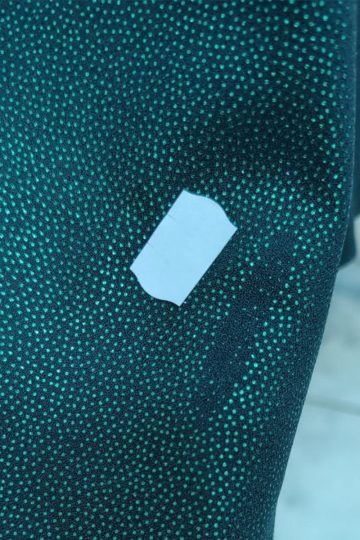 Mała biała naklejka na ciemnozielonej tkaninie w wzór drobnych jasnozielonych kropek, przypominająca elegancką zieloną brokatową sukienkę znalezioną w ekskluzywnym sklepie outletowym.