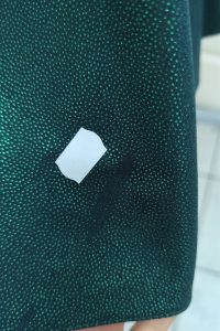Biała naklejka na ciemnozielonej tkaninie w kropki, przypominająca „Zieloną brokatową sukienkę”, z małą czarną plamką obok naklejki — idealna na znaleziska z outletu.