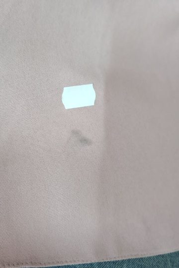 Kawałek jasnobrązowej tkaniny, przypominający sukienkę typu maxi, z białą metką i małą ciemną plamką pod nią.