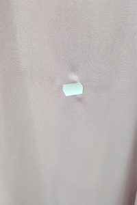 Na jasnoróżowej tkaninie, pod którą widoczne jest niewielkie wgłębienie, przyklejono mały kawałek białej taśmy, co przypomina marszczenie w talii, często spotykane w projektach sukienek typu maxi.