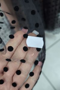 W dłoni trzyma się czarną siateczkę w białe kropki z przymocowaną do niej małą białą metką – idealną do stworzenia stylowej sukienki z długim rękawem lub znalezienia inspiracji do kolejnej stylizacji z wyprzedaży sukienek.