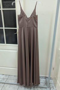 Długa, bezrękawowa sukienka satynowa w kolorze cappuccino wisi na przezroczystym wieszaku przed białymi drzwiami; jej subtelne rozcięcie na nodze dodaje jej elegancji.